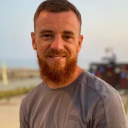 Portret uśmiechniętego mężczyzny z rudą brodą w szarej koszulce na rozmytym tle plaży i budynku.