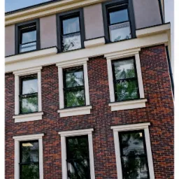 Fasada budynku z cegły z rzędami okien w czarnych ramach i białych obramowaniach, odbicie drzew w szybach, widok z dołu.