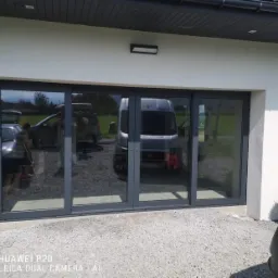 Szare, czteroskrzydłowe drzwi przesuwne z ciemnymi szybami w nowoczesnym domu z jasną elewacją i szarą okładziną na narożniku, widoczne odbicia samochodów i krajobrazu.