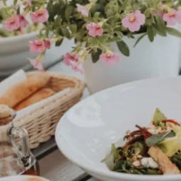 Estetyczne ujęcie stołu z białymi naczyniami, sałatką z rukolą, burakiem i serem, pieczywem w wiklinowym koszyku, kwiatami w białej doniczce oraz butelką oliwy z korkiem.