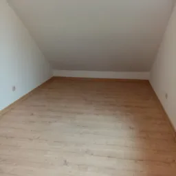 Poddasze z jasnymi panelami podłogowymi, białe ściany i skośny sufit, widoczne gniazdko elektryczne.