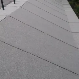 Dach pokryty papą termozgrzewalną z widocznymi pionowymi elementami wystającymi ponad powierzchnię dachu, na tle zieleni drzew i częściowo widocznego budynku z czerwoną dachówką.