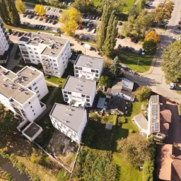 Widok z lotu ptaka na osiedle mieszkaniowe z budynkami o płaskich dachach i panelami słonecznymi na jednym z domów, otoczone zielenią i parkingami w słoneczny dzień.