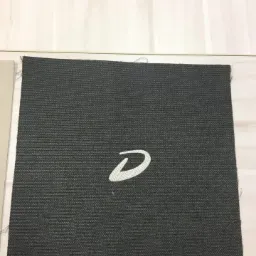 Kawałek szarej tkaniny o wyraźnej fakturze z wyhaftowanym białym, stylizowanym motywem graficznym w kształcie litery 'D', leżący na jasnym, drewnianym podłożu.