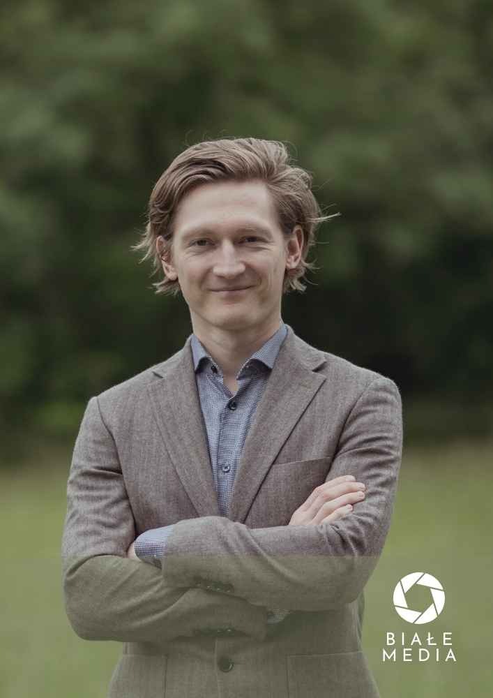Portret uśmiechniętego mężczyzny w tweedowej marynarce na rozmytym zielonym tle, z logo firmy Białe Media w prawym dolnym rogu.