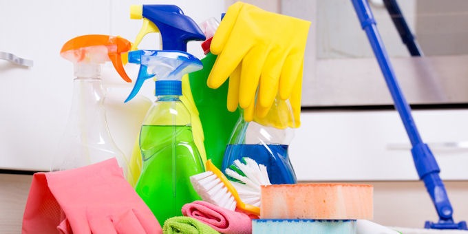 Zestaw środków czystości: kolorowe butelki ze spryskiwaczami, gąbki, szczotki, ściereczki i gumowe rękawice w odcieniach różu i żółci, ustawione na tle białych szafek.