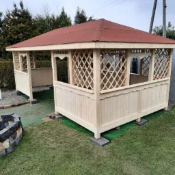 Surowa drewniana konstrukcja altany z czerwoną dachówką, widoczna kratownica i wykończenia, umieszczona na trawniku z elementami imitacji trawy, w tle drzewa i słup energetyczny.