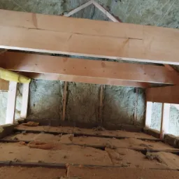 Poddasze w trakcie prac izolacyjnych, widoczne drewniane belki konstrukcyjne i częściowo ułożona warstwa izolacji z wełny mineralnej, zabezpieczona sznurkami.