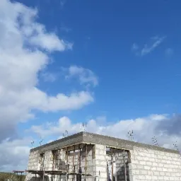 Niski budynek z betonu komórkowego w trakcie budowy, widoczne szalunki i wystające pręty zbrojeniowe na tle błękitnego nieba z chmurami.