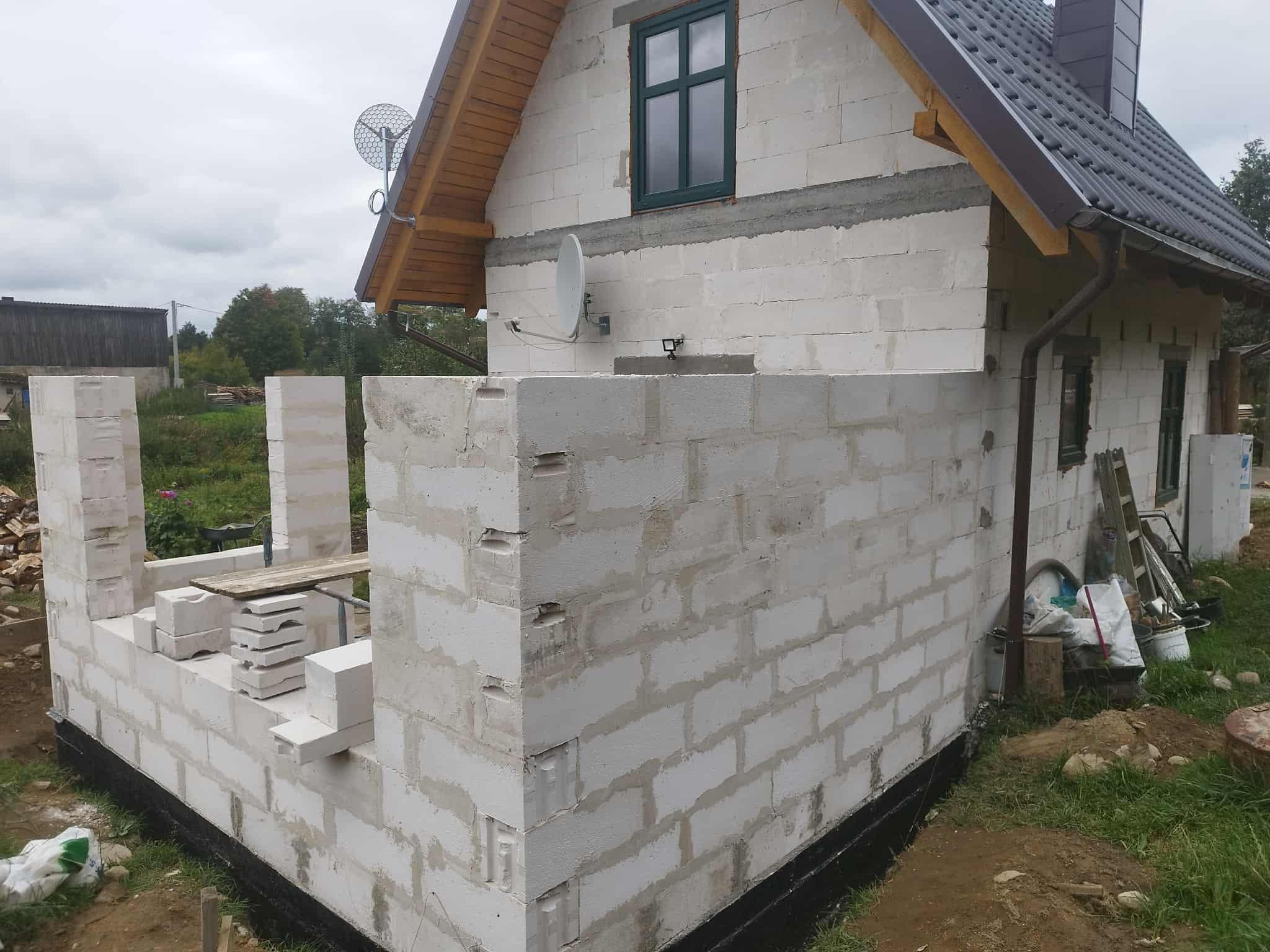 Budowa domu z białych bloczków: wznoszenie ścian parteru, widoczne zbrojenia i przygotowane materiały budowlane. Na ścianie zamontowana antena satelitarna.