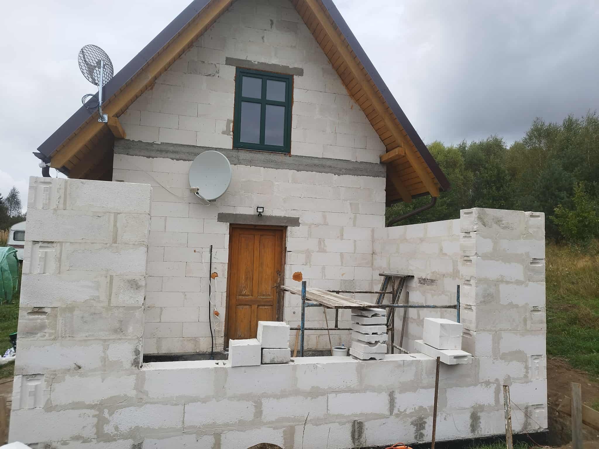 Niedokończony dom z białych bloczków, widoczne rusztowanie, drewniane drzwi i okno z zieloną ramą. Na dachu antena satelitarna.