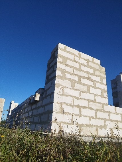 Wznoszenie ścian domu z białych bloczków betonu komórkowego, widoczne spoiny zaprawy. Budowa w toku na tle błękitnego nieba i zielonej trawy.