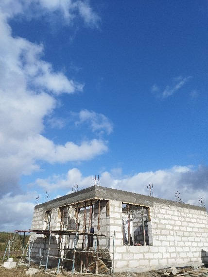 Niski budynek z betonu komórkowego w trakcie budowy, widoczne szalunki i wystające pręty zbrojeniowe na tle błękitnego nieba z chmurami.