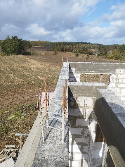 Budowa domu z betonu komórkowego: ściany wzniesione, zbrojenie wystaje ponad mur. Widok z góry na konstrukcję i okoliczne pola w słoneczny dzień.