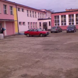 Dwupiętrowy budynek pomalowany na różowo z żółtym fragmentem, trzy samochody na placu, osoba w ciemnym ubraniu przy wejściu, betonowy plac z widocznymi spękaniami i naprawami.