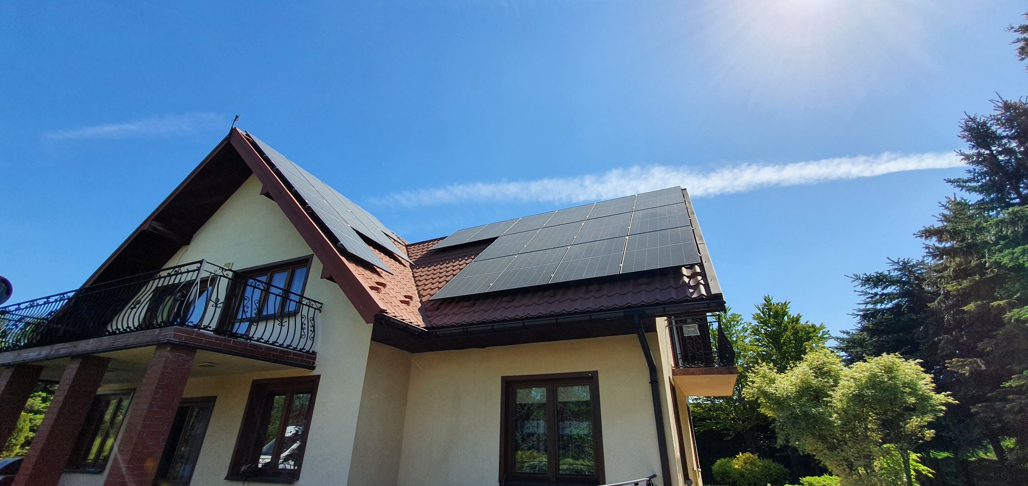 Instalacja fotowoltaiczna na dachu dwuspadowym domu jednorodzinnego, widok z dołu na panele i błękitne niebo z lekką smugą kondensacyjną.