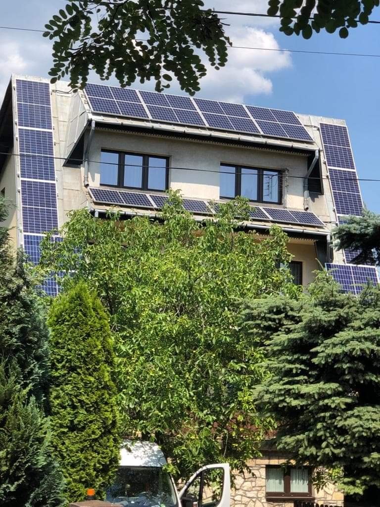 Dom jednorodzinny z panelami słonecznymi zamontowanymi na dachu i elewacji, częściowo zasłonięty zielenią drzew i krzewów, z fragmentem białego samochodu dostawczego widocznym w dolnej części kadru.