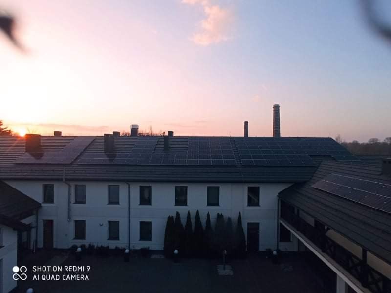 Duży budynek z panelami słonecznymi na dachu, widok z góry o zachodzie słońca, kominy w tle.