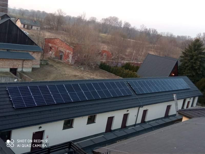 Instalacja paneli słonecznych na dachu budynku z kilkoma drzwiami wejściowymi, widok z góry, w tle zabudowania i drzewa.