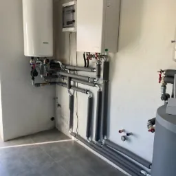 Instalacja grzewcza w kotłowni: biały zasobnik ciepłej wody, biały kocioł wiszący z osprzętem, rury w szarej izolacji, szara podłoga z płytek.