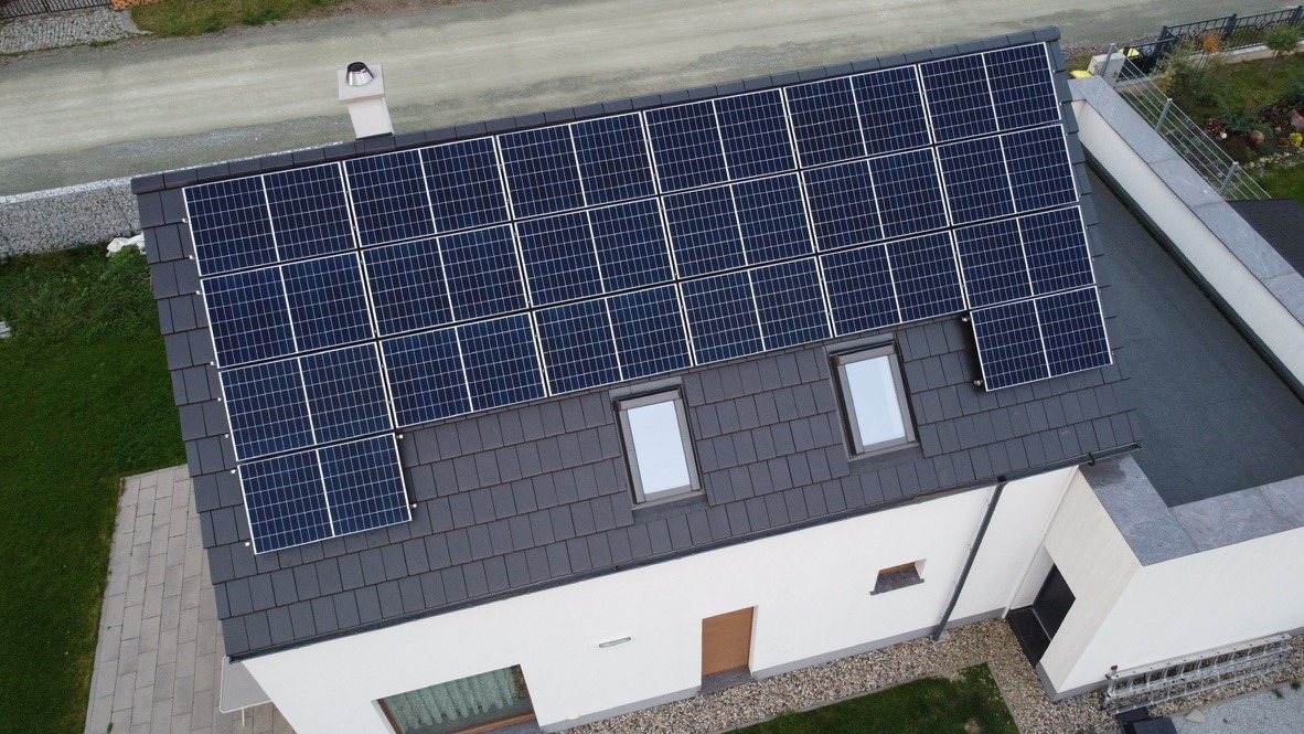 Widok z góry na dach domu z panelami słonecznymi, dwoma oknami dachowymi i kominem, otoczony zielenią.