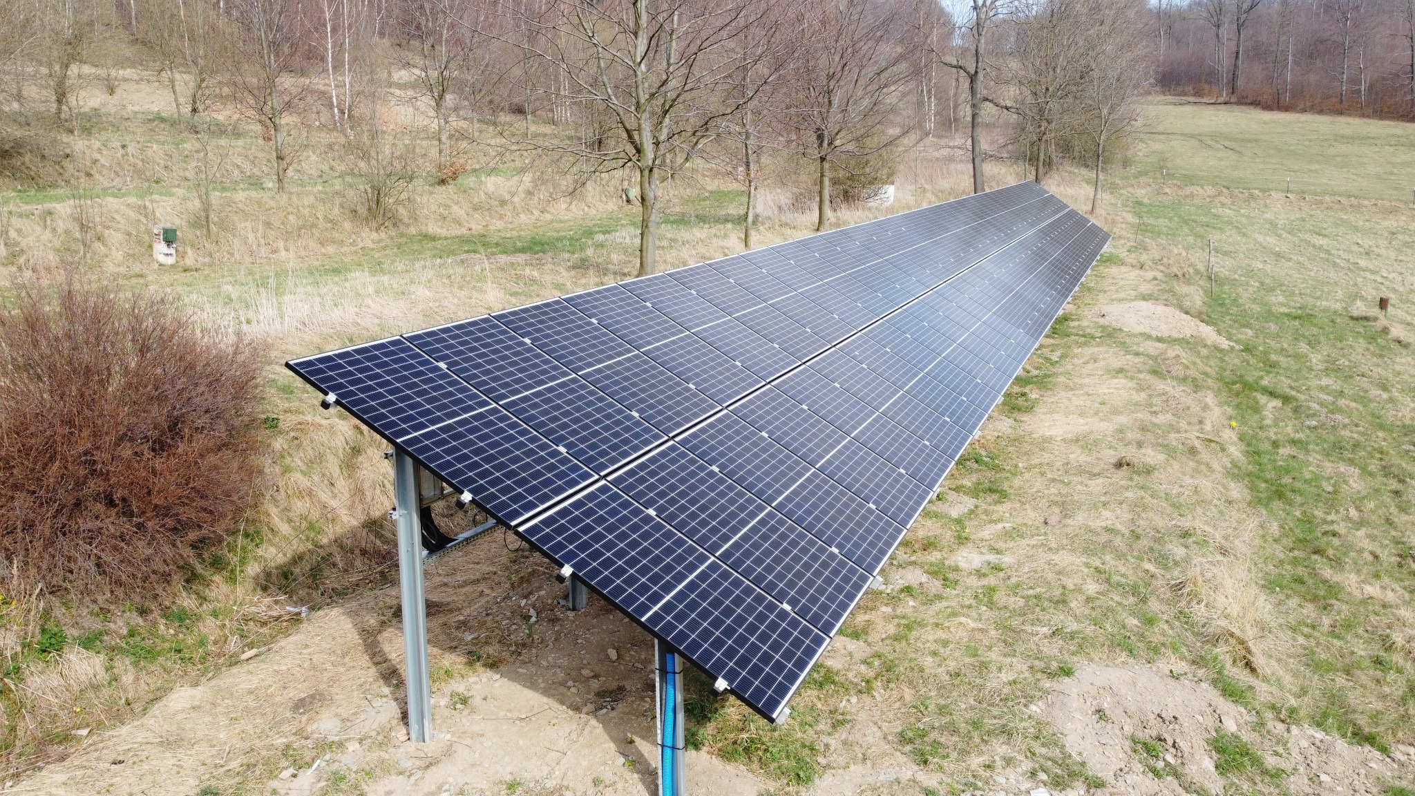 Długi rząd paneli słonecznych na metalowej konstrukcji, zainstalowany na wzgórzu z suchą trawą i młodymi drzewami w tle.
