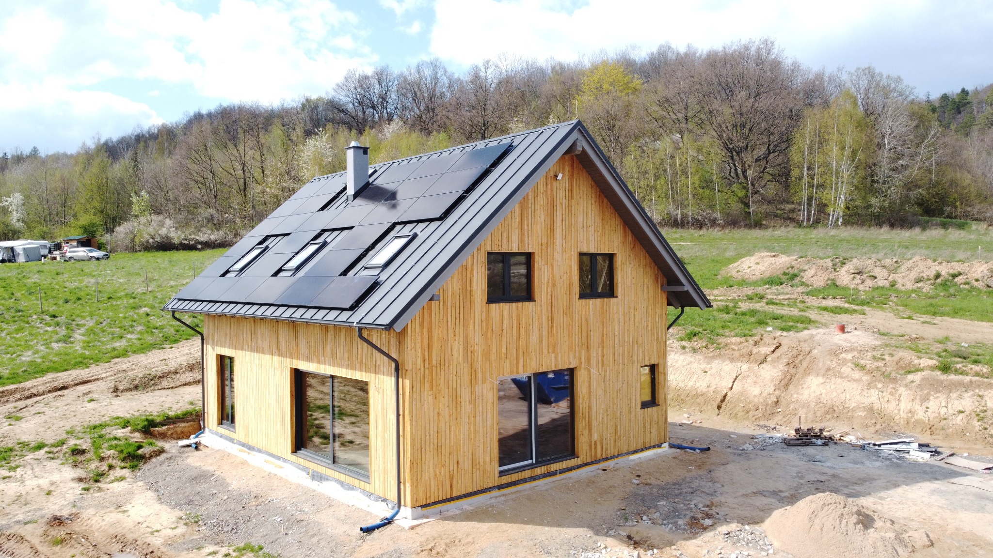 Nowoczesny dom z drewnianą elewacją i panelami słonecznymi na dachu, widok z boku na tle zieleni i drzew.