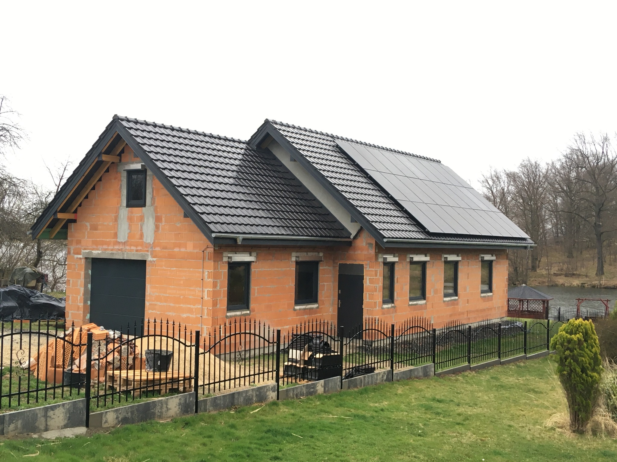 Dom w trakcie budowy z zainstalowanymi panelami fotowoltaicznymi na dachu, widoczne cegły, czarna dachówka i ogrodzenie, w tle jezioro i altana.