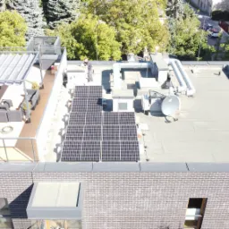 Widok z góry na dach budynku z zainstalowanymi panelami słonecznymi, anteną satelitarną i elementami wentylacji; na dachu znajdują się również dwie osoby, a w tle widoczne są drzewa i inne budynki.