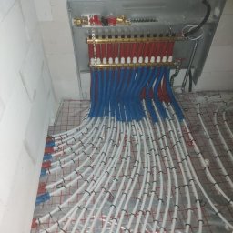 ESOLARE Sp. z o. o. - Rozdzielacz ogrzewania podłogowego z rurami w kolorach czerwonym, niebieskim i białym, ułożonymi na folii z nadrukiem siatki, zamocowanymi czarnymi uchwytami.