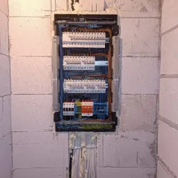 Rozdzielnica elektryczna zamontowana w ścianie z betonu komórkowego, widoczne kable i zabezpieczenia. Instalacja w trakcie prac wykończeniowych.
