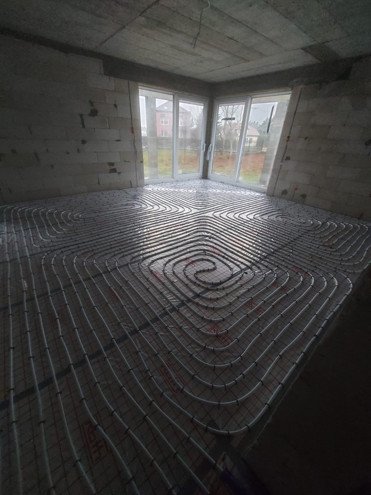 Wnętrze w surowym stanie z rozłożonym systemem ogrzewania podłogowego, widoczne rury w kształcie spiral na folii izolacyjnej, okno z widokiem na dom.