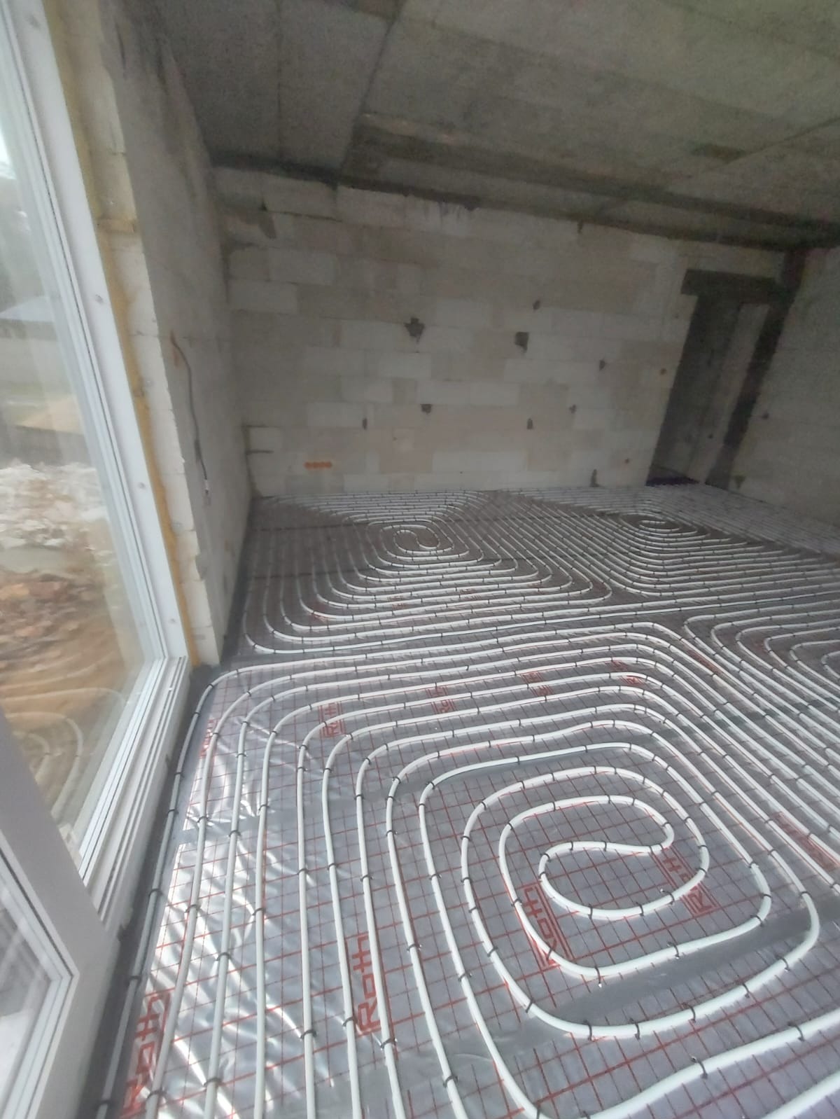 Białe rury ogrzewania podłogowego ułożone spiralnie na folii z siatką w surowym pomieszczeniu z oknem i ścianami z bloczków. Widoczny etap instalacji.