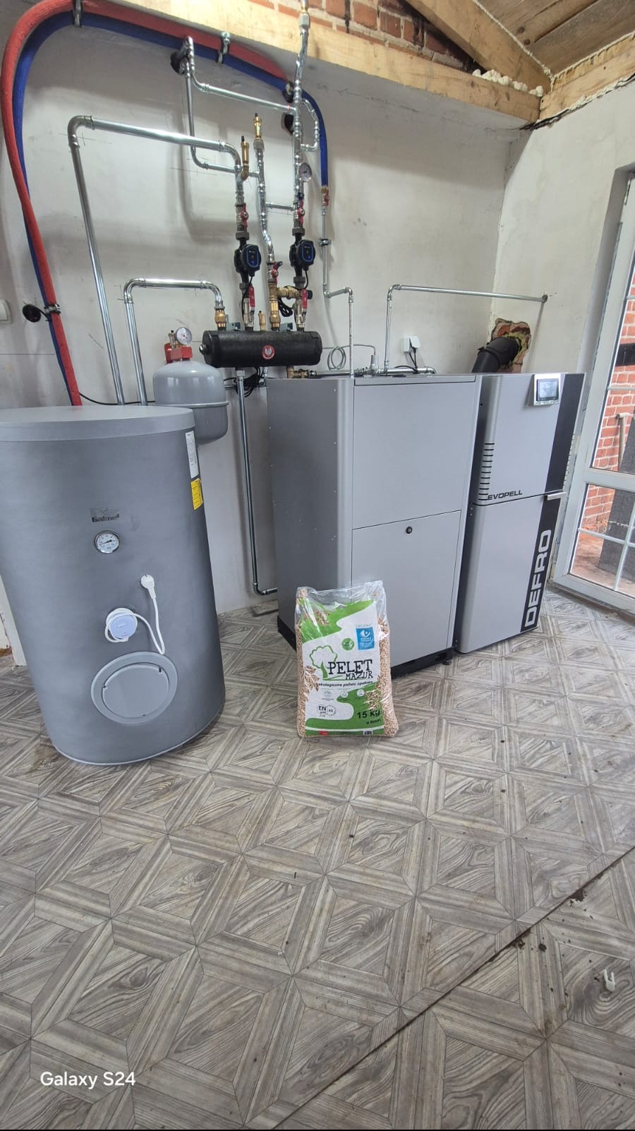 Nowoczesny piec na pellet Defro Evopell, zbiornik Galmet i instalacja grzewcza z rurami miedzianymi. W tle ceglana ściana i drewniany strop.