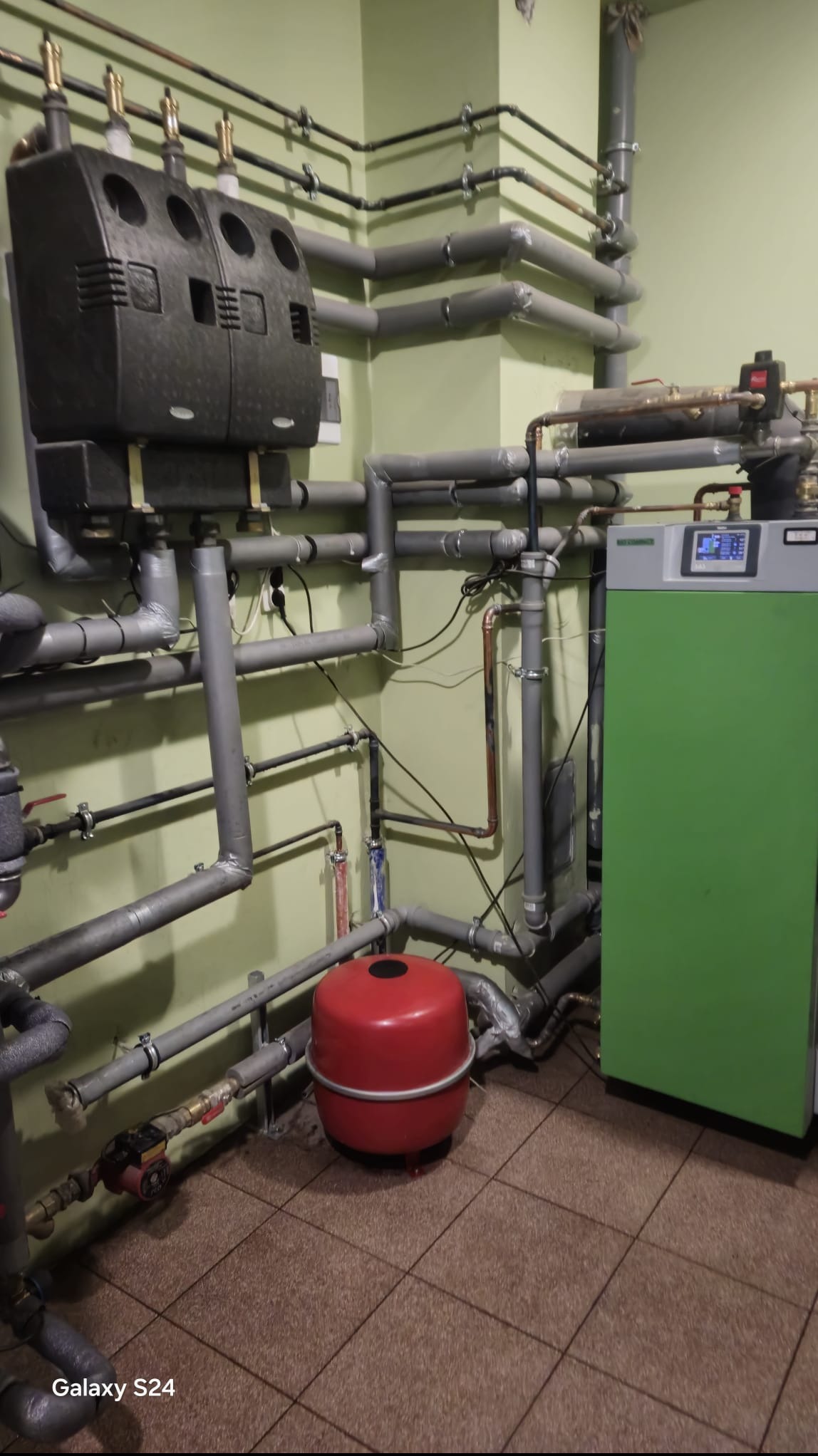 Złożona instalacja grzewcza z kotłem, rurami i czerwonym zbiornikiem wyrównawczym w pomieszczeniu o zielonych ścianach i brązowej podłodze.