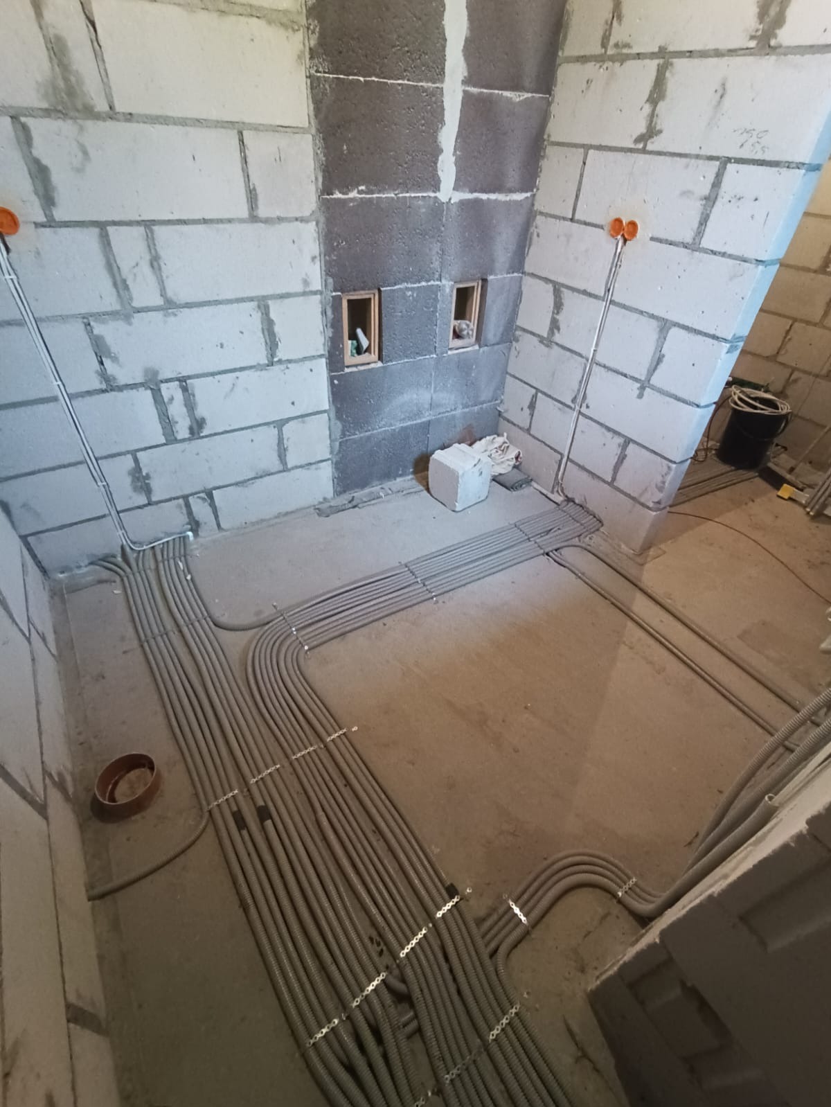 Szare rury instalacji elektrycznej ułożone równolegle na podłodze w surowym wnętrzu z bloków. Widoczne puszki elektryczne w ścianach i otwory wentylacyjne.