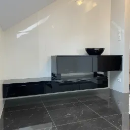 Czarna, lakierowana szafka RTV z szufladami i półkami, umieszczona na tle białych, błyszczących płytek ściennych w skosie poddasza, z czarną misą dekoracyjną na blacie.