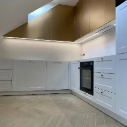 Nowoczesna kuchnia z białymi szafkami dolnymi i złotymi szafkami górnymi pod skosem dachu, oświetlenie LED podszafkowe, parkiet ułożony w jodełkę.