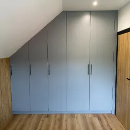 Szara zabudowa szafy z frontami bez uchwytów, dopasowana do skosu dachu, z drewnianą okładziną ściany obok i podłogą z paneli imitujących drewno.