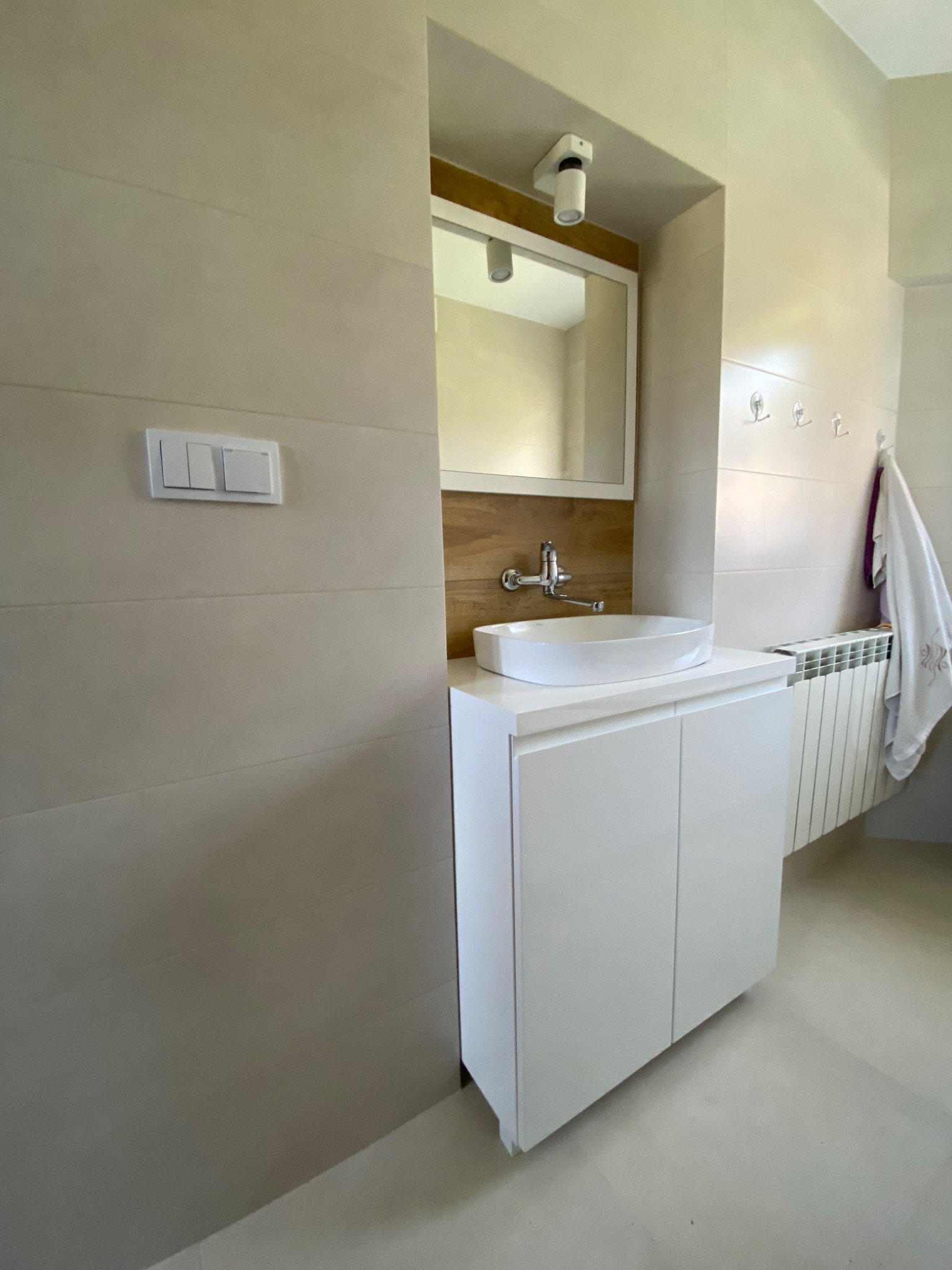 Biała szafka łazienkowa pod umywalkę z frezowanymi uchwytami, lustro w drewnianej ramie, chromowana bateria, całość w łazience wykończonej beżowymi płytkami.