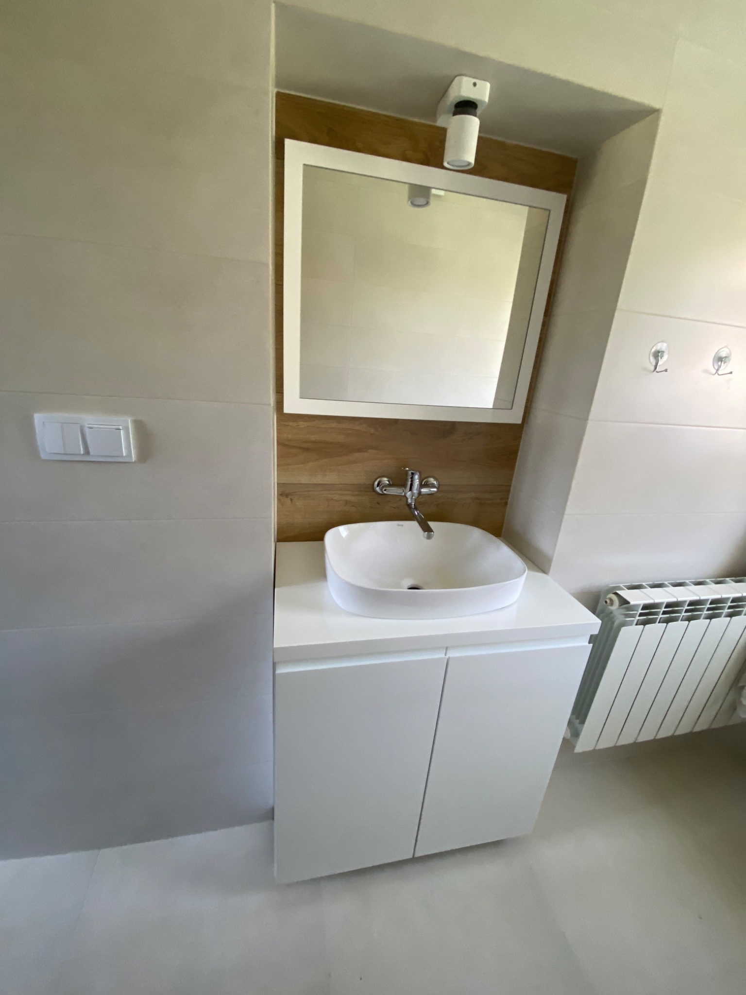 Biała szafka umywalkowa z frezowanym uchwytem, lustro wklejone w wyfrezowaną płytę, zawieszone na niewidocznych zawieszkach, bateria umywalkowa chromowana, w tle grzejnik.