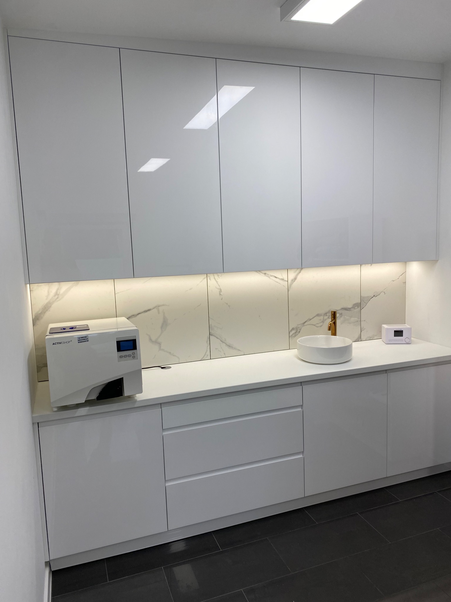 Białe, lakierowane szafki wiszące i dolne z szufladami, podświetlony blat z marmurowym wzorem, okrągła umywalka nablatowa i sprzęt medyczny na blacie.