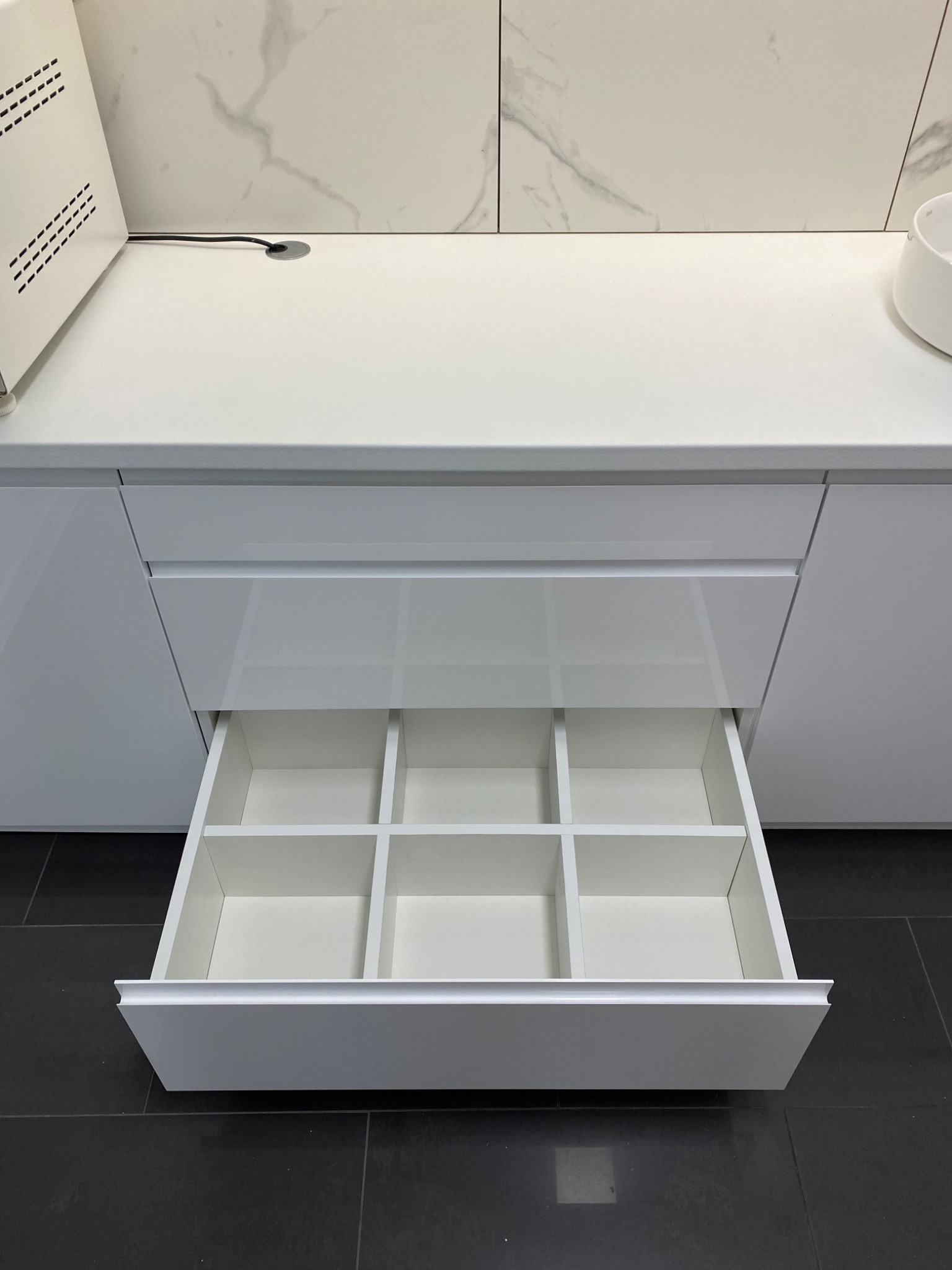 Otwarta, biała szuflada z przegródkami w nowoczesnej zabudowie kuchennej, widoczny fragment blatu i marmurowej ściany, czarna podłoga.