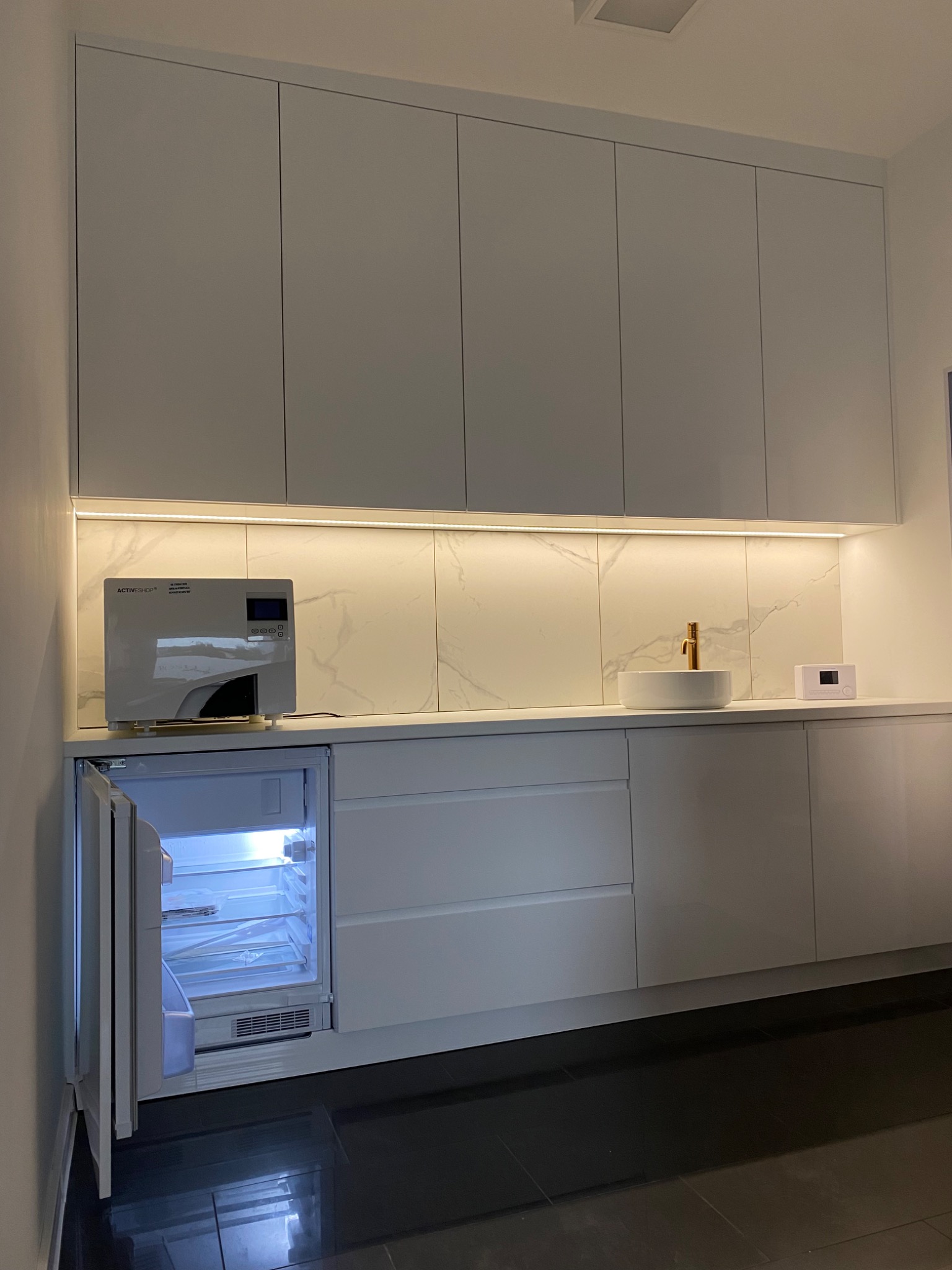 Białe szafki wiszące i dolne z oświetleniem LED pod spodem, marmurowy backsplash, okrągła umywalka z złotą baterią, otwartą chłodziarką i urządzeniem sterylizującym na blacie.