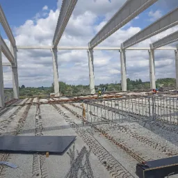 Wnętrze budowanej hali z widoczną konstrukcją stalową, zbrojeniami w betonie i pracownikami w oddali, na tle nieba z chmurami.