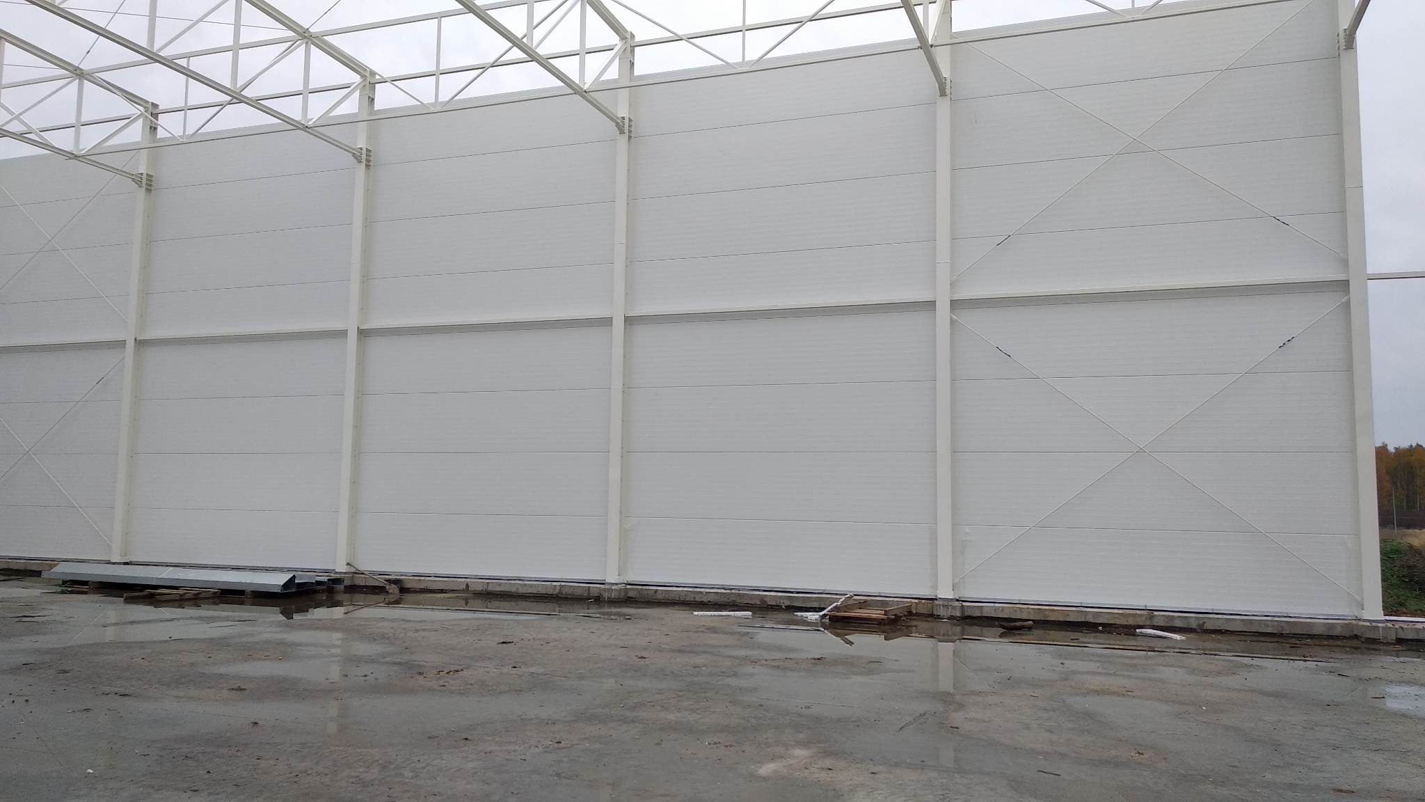Wnętrze hali w trakcie budowy, widoczne białe płyty warstwowe na ścianach i stalowa konstrukcja dachu, mokra betonowa posadzka.