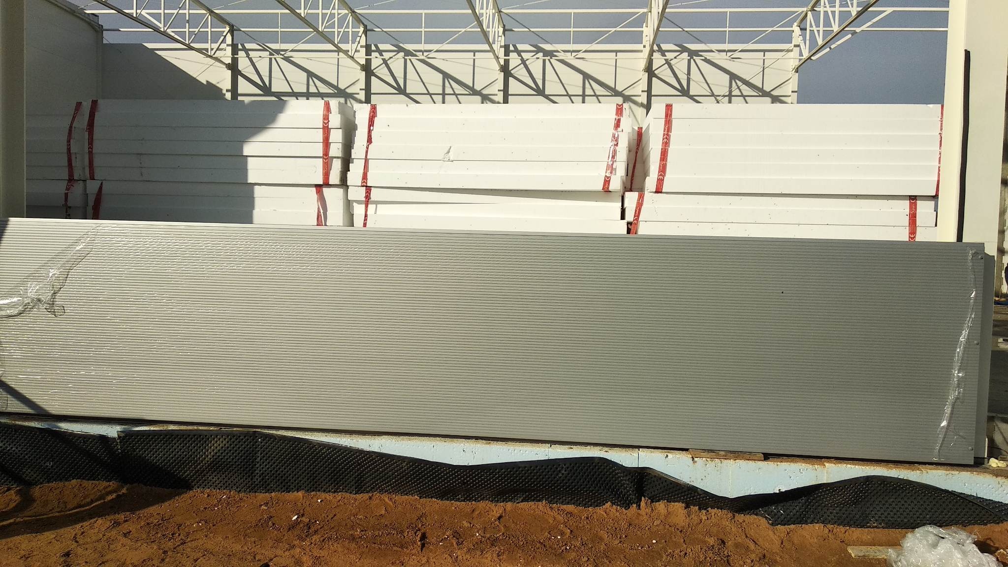 Stos płyt warstwowych zabezpieczonych folią i taśmą, składowanych na placu budowy hali stalowej z widoczną konstrukcją dachu.