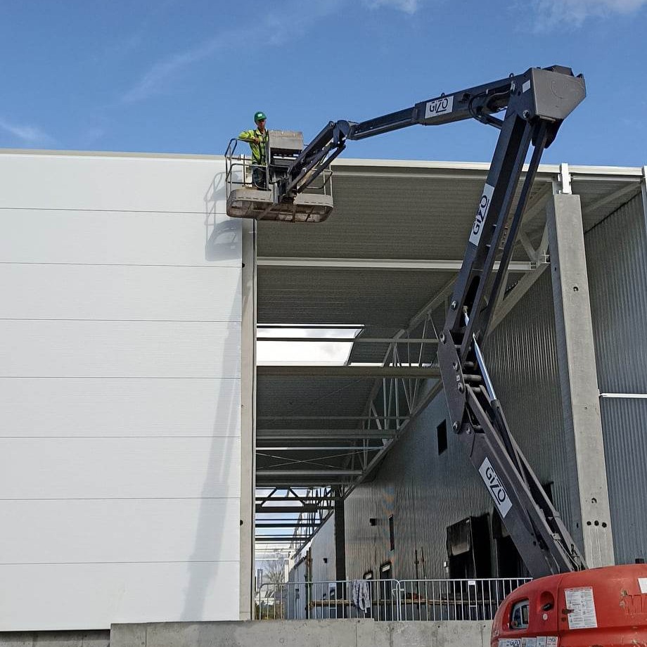 Pracownik w koszu podnośnika Gizo montuje białe płyty warstwowe na ścianie powstającej hali w Puchaczowie. Widoczna stalowa konstrukcja budynku w tle.