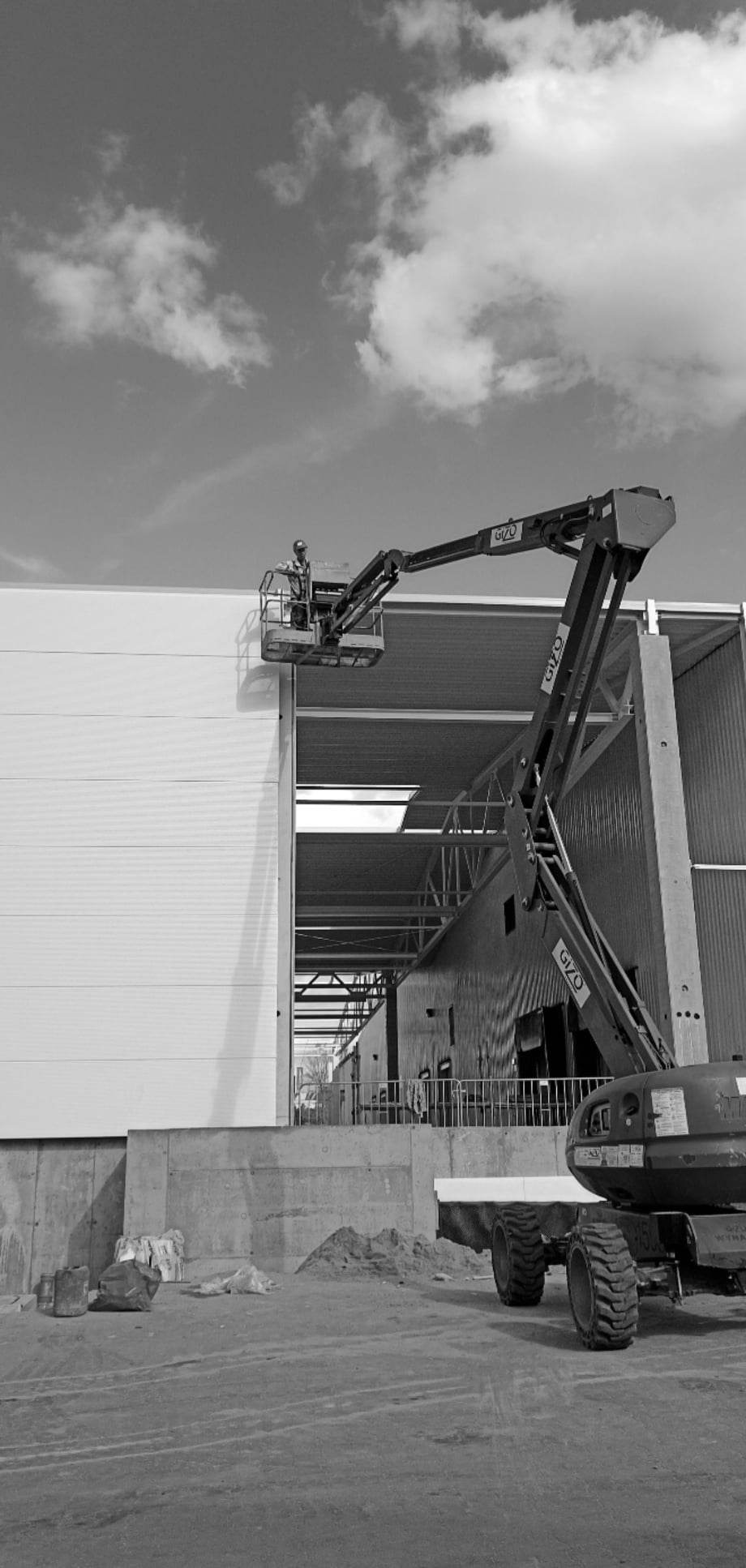 Czarno-białe zdjęcie przedstawia pracownika w koszu podnośnika, montującego białe płyty na ścianie hali. W tle widoczna stalowa konstrukcja budynku i pochmurne niebo.