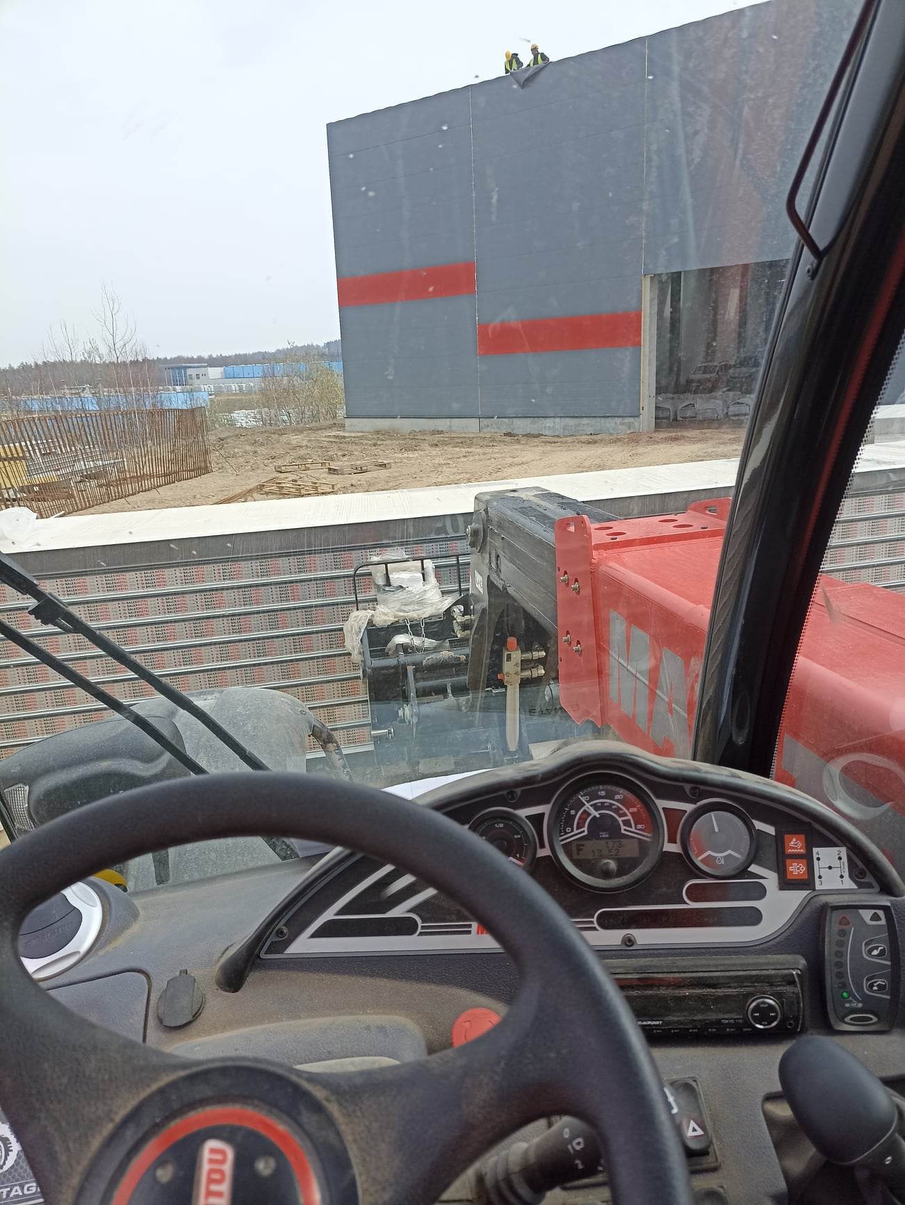 Widok z kabiny podnośnika teleskopowego na budynek hali przemysłowej z szarymi ścianami i czerwonymi pasami, gdzie dwóch pracowników pracuje na dachu.